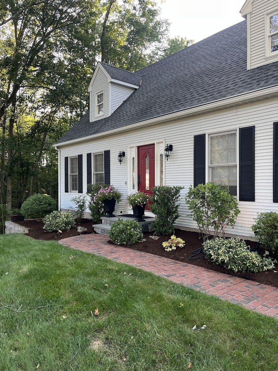 A white colonial house with a brick walk, mulched foundation shrubs, and flower pots by a red front door.